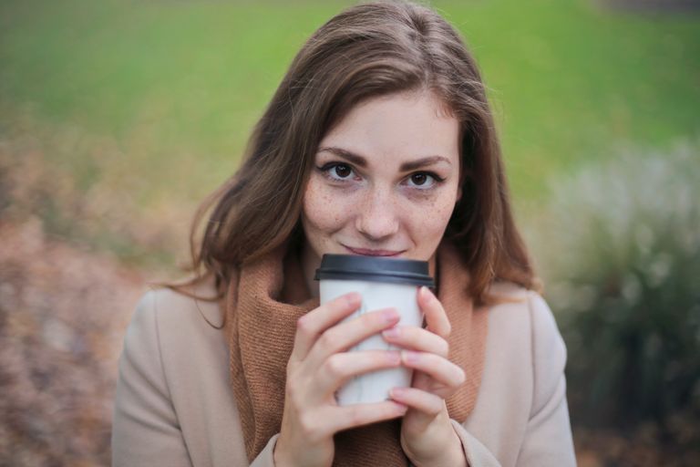 A young woman smiling while holding a coffee cup in a park during fall.
