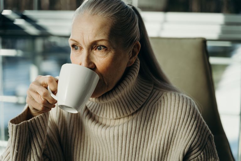 Elderly woman savoring a hot beverage indoors, in cozy attire.
