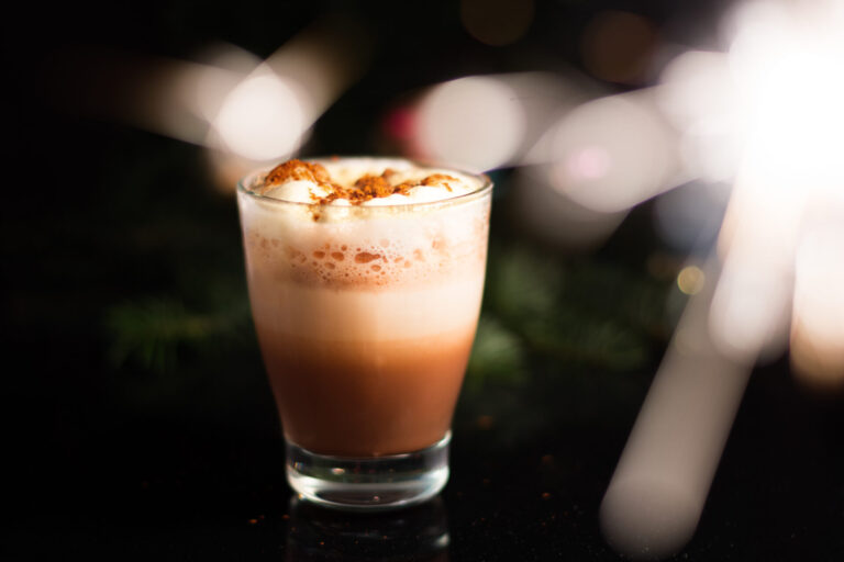Chocolate drink topped with froth and cocoa powder against warm festive lights, illustrating the chocolate base for a decaf peppermint mocha